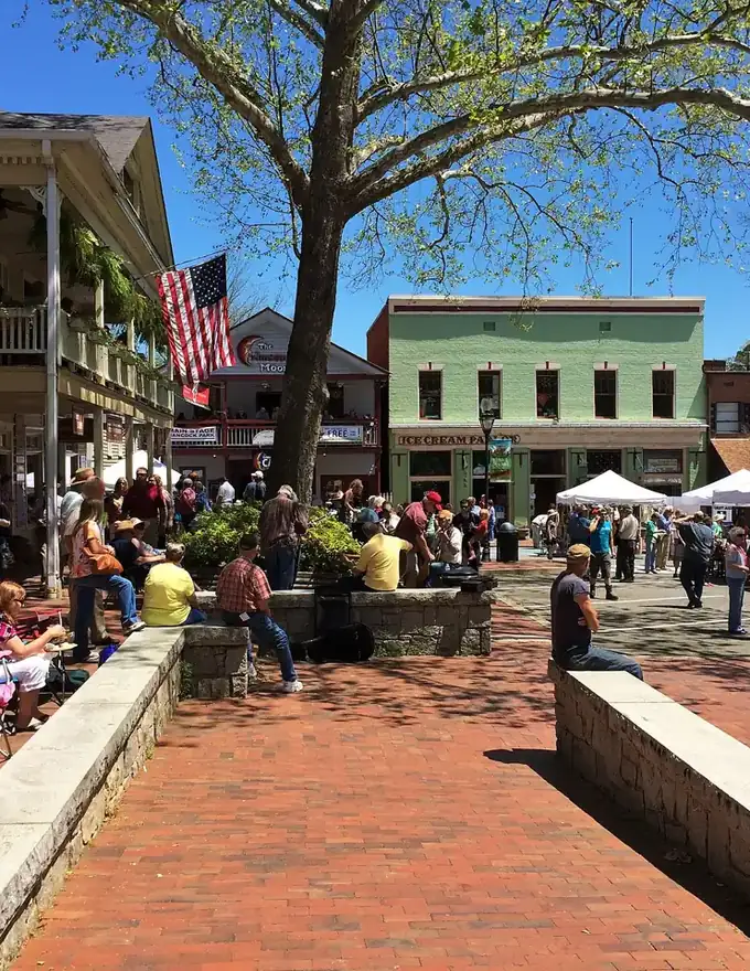 Brick plaza and historic storefronts on the Dahlonega public square, with an American flag, a large shade tree and people seated along low stone walls
