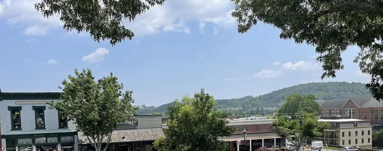 Historic storefronts on the Dahlonega public square under shade trees, with a wooded hill beyond, seen from the Gold Museum