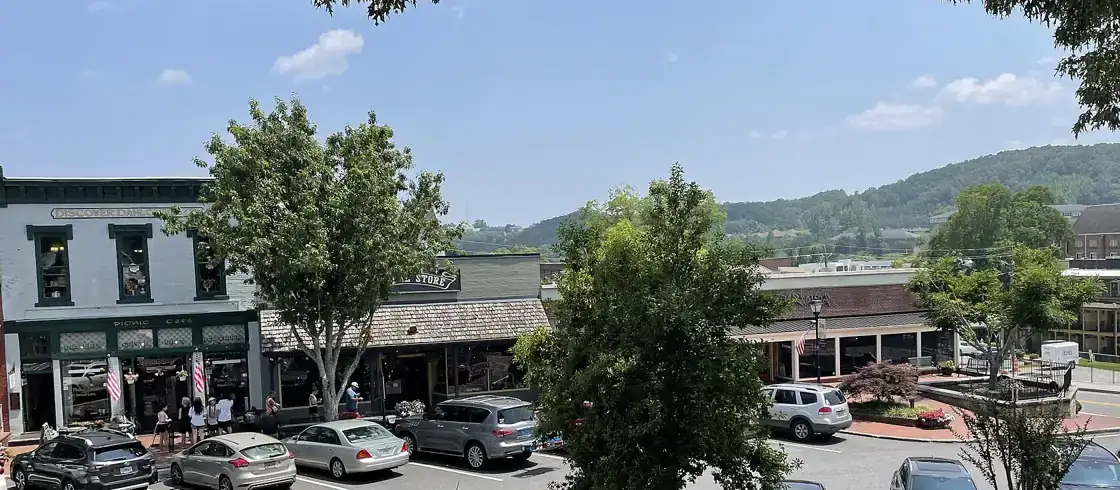 Historic storefronts and parked cars on the Dahlonega public square under shade trees, with a wooded hill beyond, seen from the Gold Museum